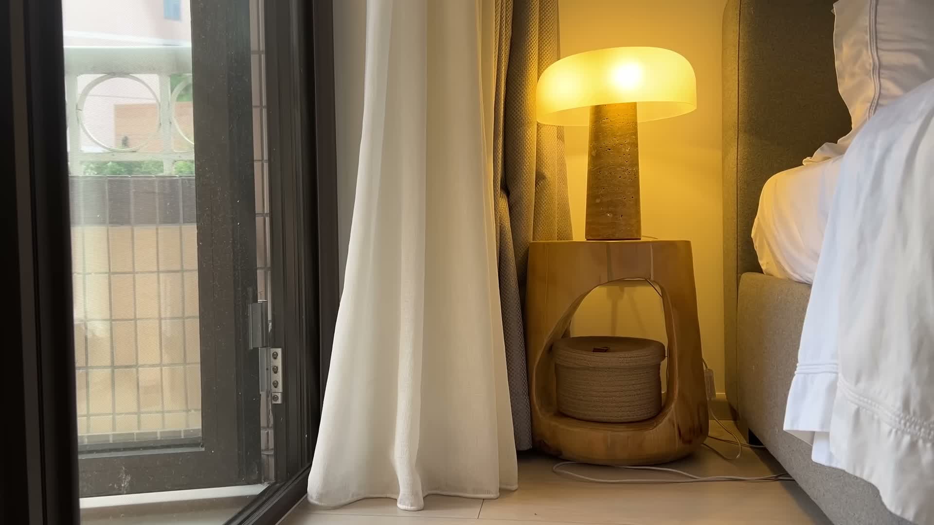 Off-white sheers diffusing daylight next to a lit mushroom lamp by the bed