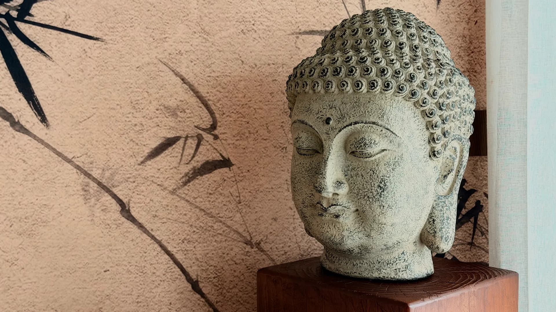 A carved stone Buddha head on a wooden plinth against a bamboo-painted wall