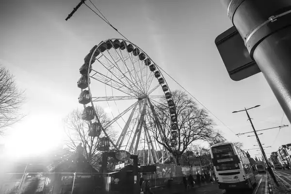 Black-and-white photograph of an Edinburgh Ferris wheel, bare trees and a bus in the low sun